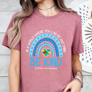 Może przedstawiać: R&oacute;żowy T-shirt z niebieską tęczą i sercem w kształcie puzzli. Tekst brzmi "W świecie, w kt&oacute;rym możesz być wszystkim, bądź miły" i "Świadomość autyzmu".