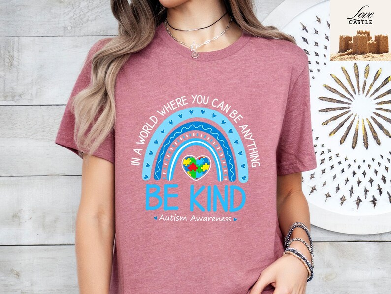 Może przedstawiać: R&oacute;żowy T-shirt z niebieską tęczą i sercem w kształcie puzzli. Tekst brzmi "W świecie, w kt&oacute;rym możesz być wszystkim, bądź miły" i "Świadomość autyzmu".