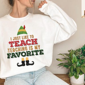 May include: White sweatshirt with the text "I JUST LIKE TO TEACH TEACHING IS MY FAVORITE" in red, green, and black. The design includes an elf hat and shoes. A fun, festive top for teachers.