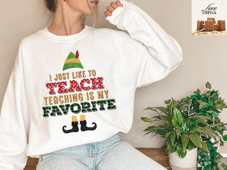 May include: White sweatshirt with the text "I JUST LIKE TO TEACH TEACHING IS MY FAVORITE" in red, green, and black. The design includes an elf hat and shoes. A fun, festive top for teachers.