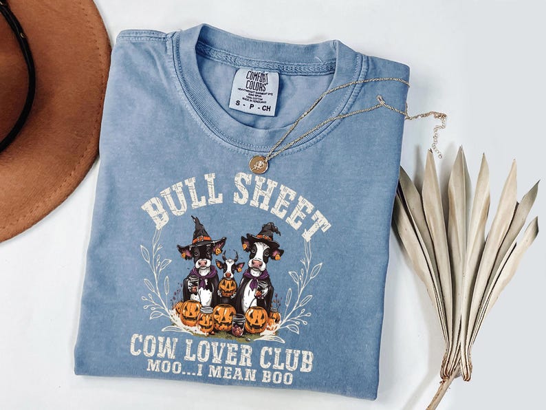 May include: A blue short-sleeve t-shirt featuring a graphic of three cows wearing witch hats surrounded by pumpkins. The text reads BULL SHEET, COW LOVER CLUB, and MOO...I MEAN BOO. The shirt has a Comfort Colors label in the neck area.