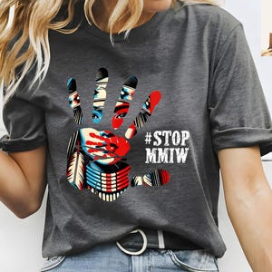 May include: Gray t-shirt with a graphic of a handprint with a Native American woman's face inside. The handprint is red, white, and blue. The text "#STOP MMIV" is printed in white below the handprint.