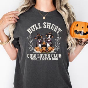 May include: A charcoal gray t-shirt featuring a graphic of three cows wearing witch hats surrounded by pumpkins. The text reads BULL SHEET COW LOVER CLUB MOO...I MEAN BOO. The design is framed by a thin white leaf wreath on the front of the shirt.
