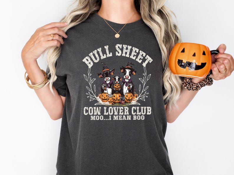 May include: A charcoal gray t-shirt featuring a graphic of three cows wearing witch hats surrounded by pumpkins. The text reads BULL SHEET COW LOVER CLUB MOO...I MEAN BOO. The design is framed by a thin white leaf wreath on the front of the shirt.
