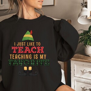 May include: Black sweatshirt with the text "I JUST LIKE TO TEACH TEACHING IS MY FAVORITE" in red, green, and white. The design includes an elf hat and shoes. A cozy and festive garment for teachers.