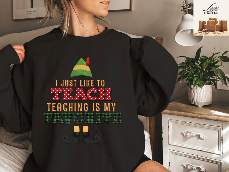 May include: Black sweatshirt with the text "I JUST LIKE TO TEACH TEACHING IS MY FAVORITE" in red, green, and white. The design includes an elf hat and shoes. A cozy and festive garment for teachers.