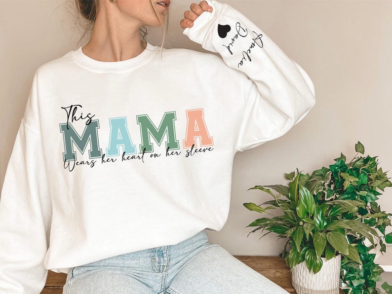 May include: White long-sleeve sweatshirt with the text "This Mama wears her heart on her sleeve" in colorful letters. The sleeve has the text "Andie" with a heart.