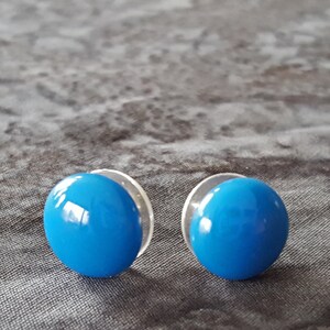 May include: A pair of round, bright blue stud earrings with a glossy finish. Each earring has a clear, circular backing. The earrings are set against a textured, dark gray background.