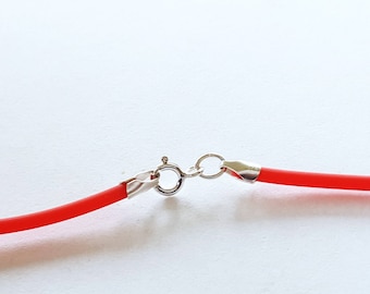 May include: Close-up of a red necklace with a silver clasp. The necklace has a bright red cord and a shiny silver clasp. The clasp is connected to the necklace with small silver rings. The background is white.