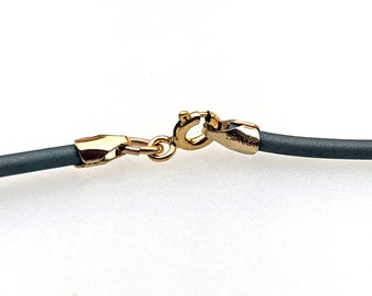 May include: Close-up of a gold-colored clasp and a dark gray cord. The clasp is connected to the cord, which appears to be part of a necklace or bracelet. The background is white.