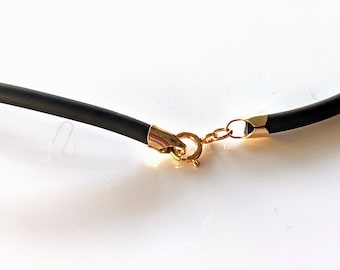 May include: Close-up of a black necklace with gold-colored clasps and accents. The necklace has a round, black cord and a gold-colored clasp to secure it. The necklace is on a white surface.