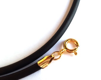 May include: Close-up of a black rubber necklace with a gold-colored clasp. The clasp is a spring ring type, and the necklace appears to be a choker style. The background is plain white, highlighting the jewelry.