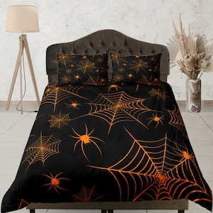 以下が含まれることがあります： 黒とオレンジの蜘蛛の巣柄の寝具セット。寝具は、オレンジ色の蜘蛛の巣と蜘蛛が描かれた黒い背景です。