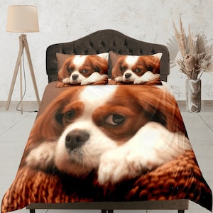 Puede incluir: Un perro Cavalier King Charles Spaniel marrón y blanco con ojos marrones está acostado sobre una manta con estampado marrón y naranja. El perro está mirando a la cámara con una expresión triste. El perro está en una cama con dos almohadas detrás. Las almohadas son blancas con un perro Cavalier King Charles Spaniel marrón y blanco impreso en ellas.
