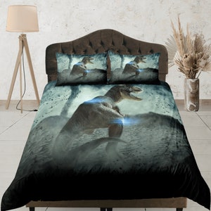 May include: A black and grey dinosaur bedding set featuring a large dinosaur graphic on the duvet cover. The dinosaur is standing on a rocky surface with a blue glow behind it. The bedding set includes two pillowcases with smaller dinosaur graphics.