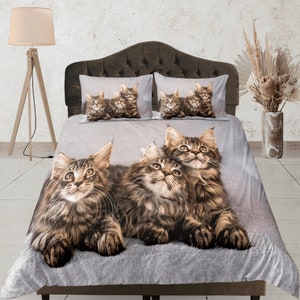 May include: A grey and white duvet cover featuring a pattern of three Maine Coon cats. The cats are brown and black with long fur. The duvet cover is on a bed with a brown headboard.