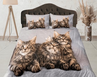 May include: A gray and white duvet cover with a pattern of three Maine Coon cats. The cats are brown and black with long fur. The duvet cover is on a bed with a brown headboard.