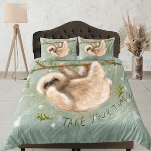 Può includere: Una federa blu-verde con un bradipo appeso a un ramo. Il bradipo è marrone e ha un'espressione carina. La federa ha un motivo a spirale bianco e oro e il testo "Take your time" in nero.