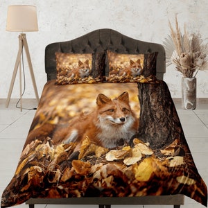 May include: A red fox sits amongst a bed of autumn leaves. The fox is looking at the camera. The bedding is brown and orange with a tree trunk in the background.