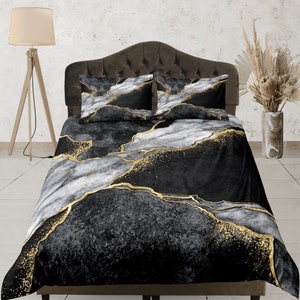 Op de afbeelding: Een zwart, wit en goudkleurig marmerpatroon beddengoed. De set bevat een dekbedovertrek en twee kussenslopen. Het ontwerp heeft een donkere achtergrond met witte en gouden aderen, wat een luxe uitstraling creëert. Het beddengoed wordt getoond op een bed met een bruin hoofdbord.
