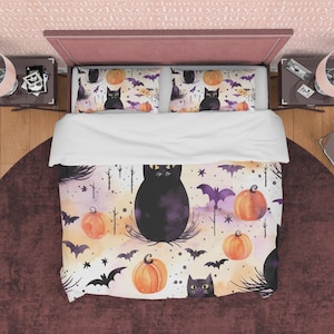 Op de afbeelding: Een witte dekbedovertrek met een Halloween-thema. De dekbedovertrek heeft een zwarte kat, vleermuizen en pompoenen op een aquarel achtergrond. De dekbedovertrek ligt op een bed met twee bijpassende kussenslopen.