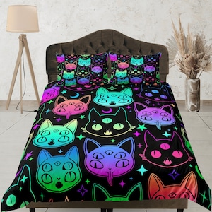 Op de afbeelding: Een zwart en regenboogkleurig dekbedovertrek met een herhalend patroon van katten met meerdere ogen. De katten zijn zwart omlijnd en hebben een regenboogkleurige verloopvulling. De achtergrond is zwart met witte sterren.