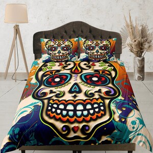 Puede incluir: Una funda nórdica colorida con un gran diseño de calavera de azúcar. La calavera es blanca con contornos negros y presenta detalles en rojo brillante, naranja, amarillo, verde, azul y morado. El fondo es una mezcla de naranja, azul y verde con patrones florales.
