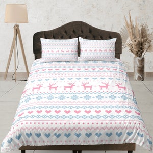 Puede incluir: Una funda nórdica blanca con un patrón escandinavo rosa y azul con renos, corazones y copos de nieve. La funda nórdica está en una cama con dos fundas de almohada a juego.