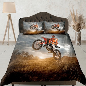 Puede incluir: Una moto de cross roja y naranja con un piloto salta sobre una colina de tierra. El fondo es un cielo nublado con una puesta de sol.