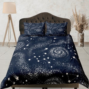 Può includere: Un completo letto blu scuro con un design celeste di stelle bianche, costellazioni e galassie vorticose. Il set include un copripiumino e due federe. La biancheria da letto è esposta su una struttura letto marrone.