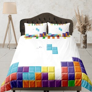 Puede incluir: Bedding blanco con un patrón de Tetris colorido en la parte inferior. La ropa de cama tiene dos fundas de almohada a juego con un patrón de Tetris.