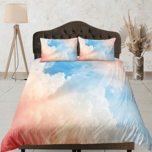 Pode incluir: Uma cama com um edredom e duas fronhas combinando com estampa de nuvens brancas, azuis e rosas. O edredom tem um design suave, semelhante à aquarela.