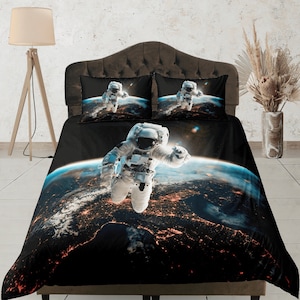 Op de afbeelding: Een zwarte dekbedovertrek met een ruimtethema met een astronaut die voor een blauwe en oranje aarde zweeft. De astronaut draagt een wit ruimtepak en een helm. De achtergrond is donkerblauw met sterren.