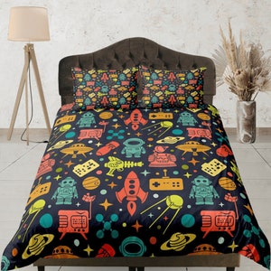 Może przedstawiać: Poszwa na kołdrę w ciemnoniebieskim kolorze z retro wzorem o tematyce kosmicznej. Wzór przedstawia roboty, statki kosmiczne, planety, gwiazdy i kontrolery do gier w odcieniach pomarańczowego, żółtego, zielonego i niebieskiego.