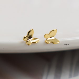 Peut inclure: Une paire de petites boucles d'oreilles à clous en forme de papillon dorés. Les boucles d'oreilles sont présentées sur une assiette blanche, avec un arrière-plan flou de perles. Le design du papillon est simple et élégant.