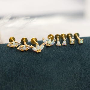May include: A collection of gold-toned earrings with various opal and crystal designs. The earrings are displayed on a dark teal surface. The image also includes a strand of white pearls and the word "coco".