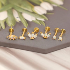 May include: A collection of gold-colored ear studs with various opal and crystal designs. The studs are displayed on a gray surface, with a blurred background of white flowers and greenery. The word "coco" is visible in the background.