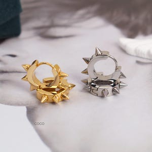 May include: Two pairs of hoop earrings with a spiked design. One pair is gold-colored, and the other is silver-colored. The earrings are displayed on a white surface with a blurred image in the background.