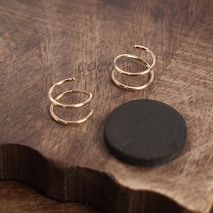May include: Two gold spiral earrings, each with two loops. They are presented on a dark brown wooden surface, accompanied by black circular objects.