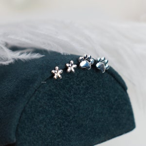 May include: Four silver earrings on a dark green surface. Two earrings are small flower shapes and two are paw print shapes.