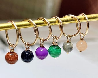 May include: A collection of gold hoop earrings, each adorned with a different gemstone bead. The beads include shades of brown, black, purple, green, gray, and peach. The earrings are displayed on a gold stand.