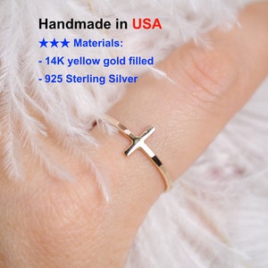 May include: A delicate, handmade ring with a cross design. The ring is made of 14K yellow gold filled or 925 Sterling Silver. The ring is shown on a finger against a soft, white background. The text "Handmade in USA" is visible.