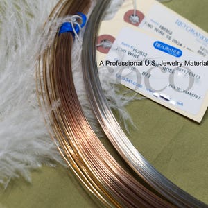 Puede incluir: Primer plano de suministros para la fabricaci&oacute;n de joyas, que incluyen carretes de alambre de color cobre, plata y oro. Tambi&eacute;n se ven una pluma blanca y una etiqueta de Rio Grande, un proveedor de materiales para joyer&iacute;a. La etiqueta dice "END WIRE SS 20GA".
