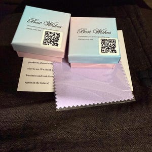 Puede incluir: Dos peque&ntilde;as cajas de regalo, una azul claro y otra rosa, con el texto "Best Wishes" y un c&oacute;digo QR. Tambi&eacute;n se ve un peque&ntilde;o pa&ntilde;o lavanda y una nota con texto.