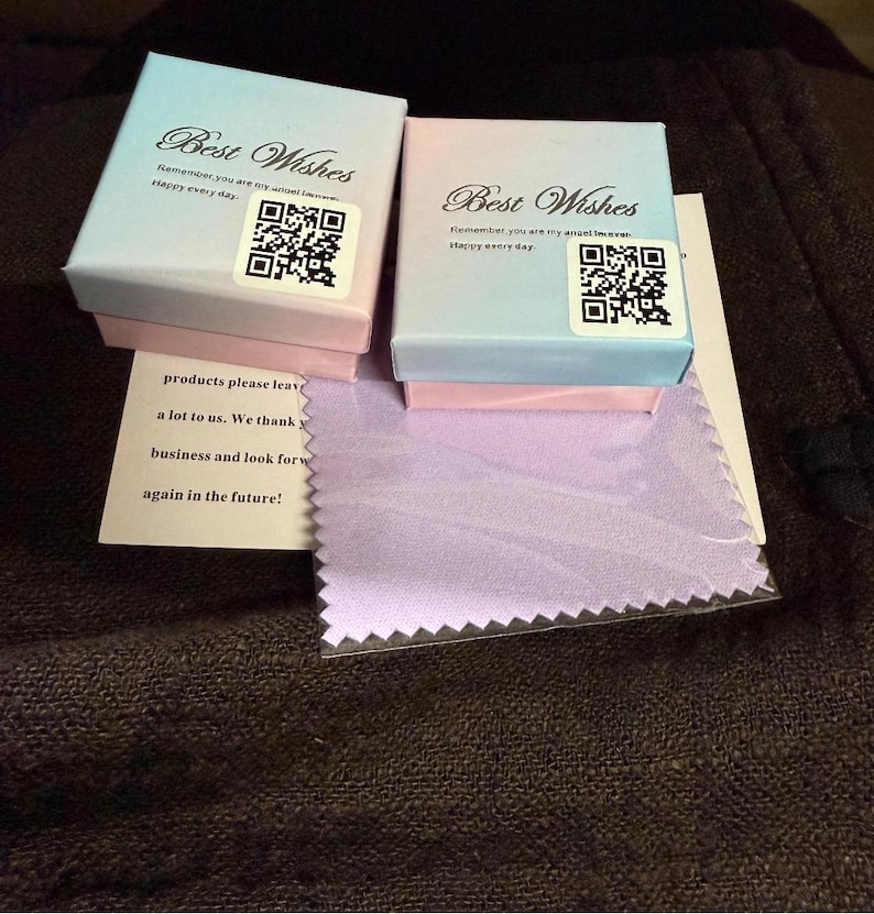 Puede incluir: Dos peque&ntilde;as cajas de regalo, una azul claro y otra rosa, con el texto "Best Wishes" y un c&oacute;digo QR. Tambi&eacute;n se ve un peque&ntilde;o pa&ntilde;o lavanda y una nota con texto.