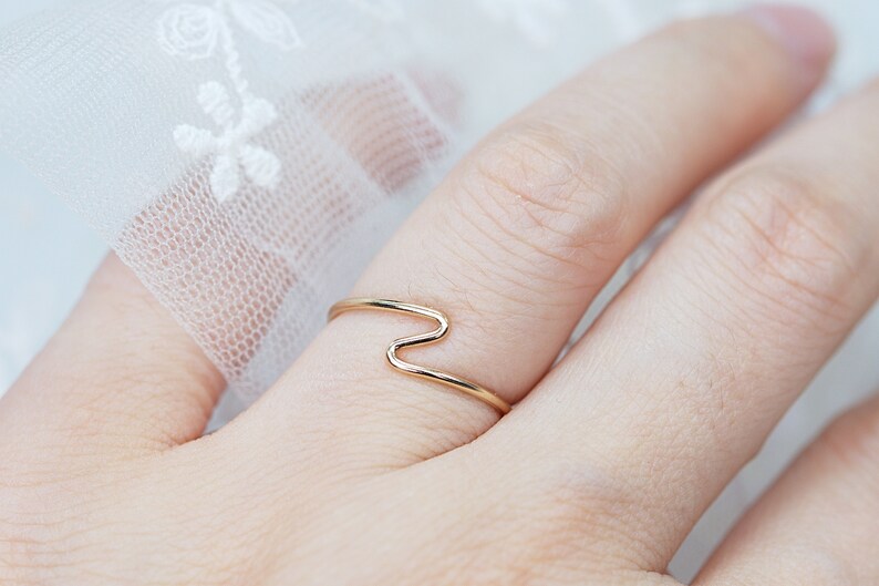 Puede incluir: Un delicado anillo de oro con un dise&ntilde;o en zigzag &uacute;nico. El anillo es fino y minimalista, con un patr&oacute;n continuo en forma de onda. La joya se muestra en un dedo, destacando su elegante simplicidad. El dise&ntilde;o del anillo es moderno y elegante.