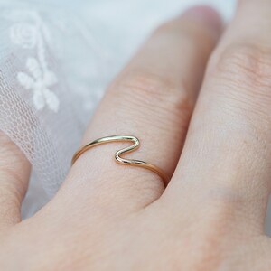 Puede incluir: Un delicado anillo de oro con un dise&ntilde;o ondulado. El anillo se lleva en un dedo, mostrando su estilo minimalista y elegante. La superficie lisa y pulida del anillo refleja la luz, resaltando sus sutiles curvas y forma.