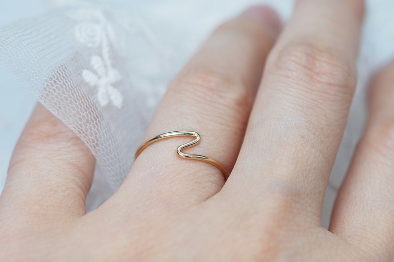 Puede incluir: Un delicado anillo de oro con un dise&ntilde;o ondulado. El anillo se lleva en un dedo, mostrando su estilo minimalista y elegante. La superficie lisa y pulida del anillo refleja la luz, resaltando sus sutiles curvas y forma.