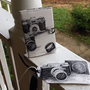 May include: A set of two items with a camera theme. One is a book-style case with a button closure, and the other is a tag. Both items feature a black and white sketch of a vintage camera.