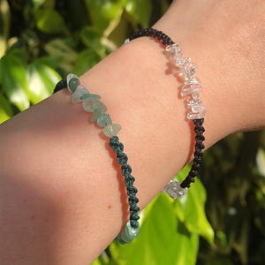 Puede incluir: Dos pulseras hechas a mano. Una presenta virutas de cristal transparente sobre un cordón negro, la otra tiene virutas de piedras preciosas verdes sobre un cordón verde oscuro. Ambas son de estilo macramé.